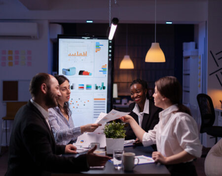 Workaholics businesspeople brainstorming financial company ideas analyzing strategy paperwork late at night in business office meeting room. Multi-ethnic coworkers checking solution